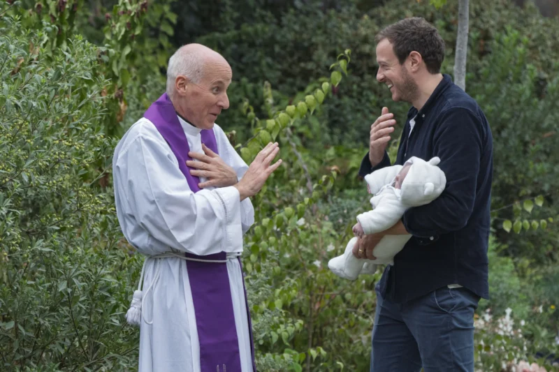 Thierry · 26/09/2026 · 3840 x 2560 Un prêtre en tenue liturgique discute avec un homme tenant un bébé, entourés de végétation dans un cadre extérieur lumineux
