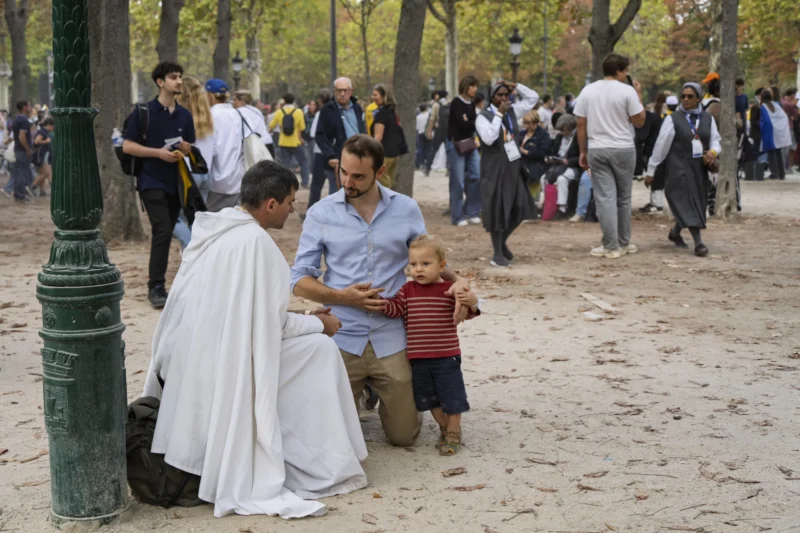 Thierry · 26/09/2026 · 3840 x 2560 Un homme en robe blanche discute avec un adulte et un enfant dans un parc animé, entouré de passants et de feuilles mortes