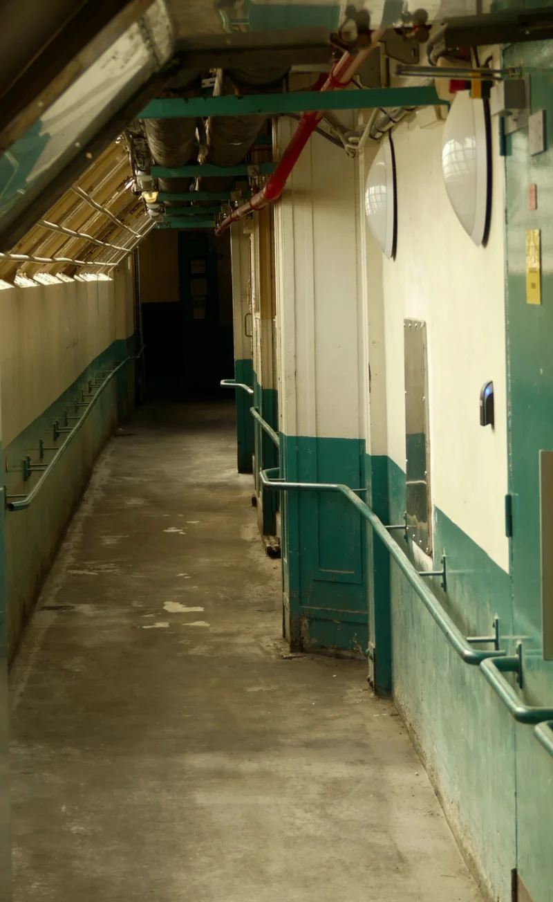 Couloir industriel avec des portes vertes, des rails métalliques et des conduits, éclairé par des néons, ambiance utilitaire