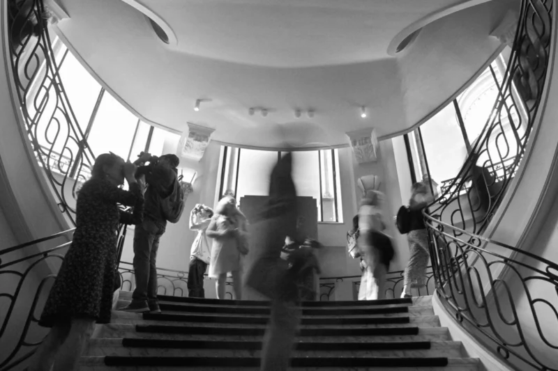 Groupe de personnes en mouvement dans un escalier intérieur lumineux avec balustrades ornées, certaines prenant des photos.