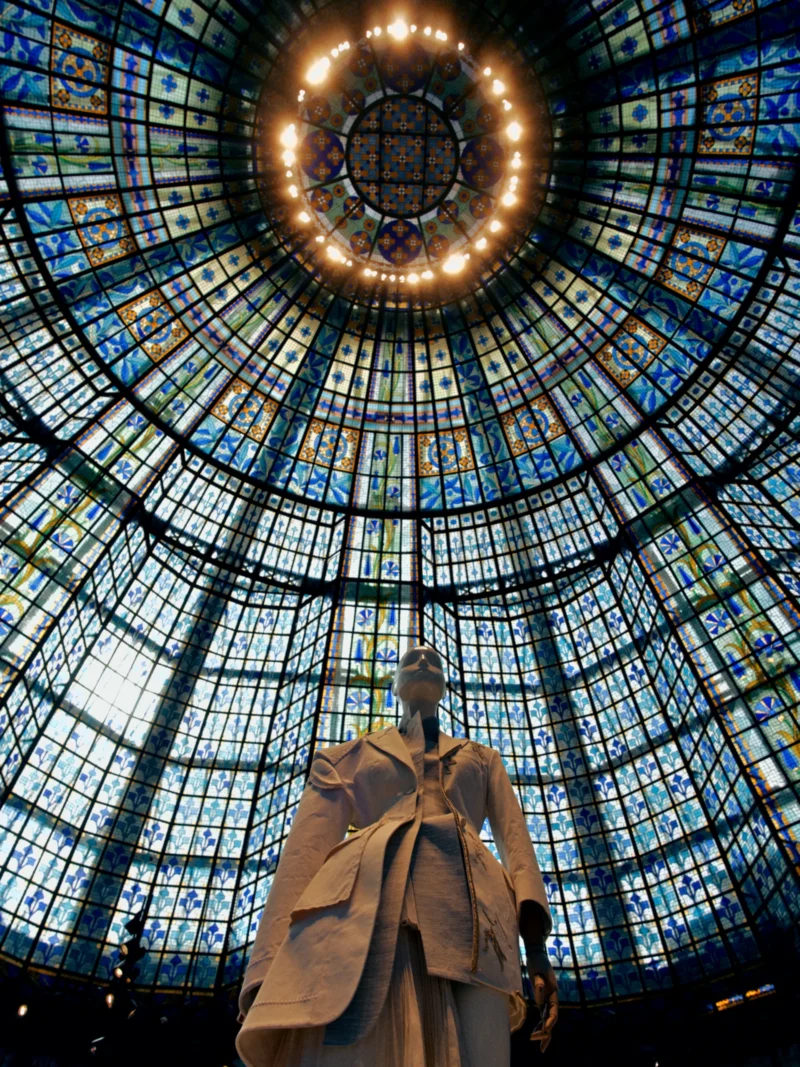 Statue vêtue d'un long manteau sous une coupole vitrée aux motifs colorés et lumineuse, vue de dessous vers le haut.
