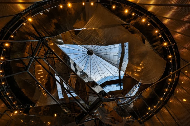 Bao · 22/08/2026 · 1536 x 1024 Vue intérieure d’un atrium moderne avec une structure métallique et une verrière éclairée par des lumières dorées en soirée