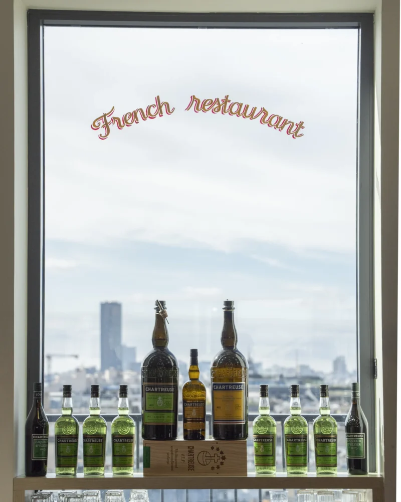 Étagère avec bouteilles de Chartreuse devant une fenêtre offrant une vue urbaine, avec enseigne 'French restaurant'