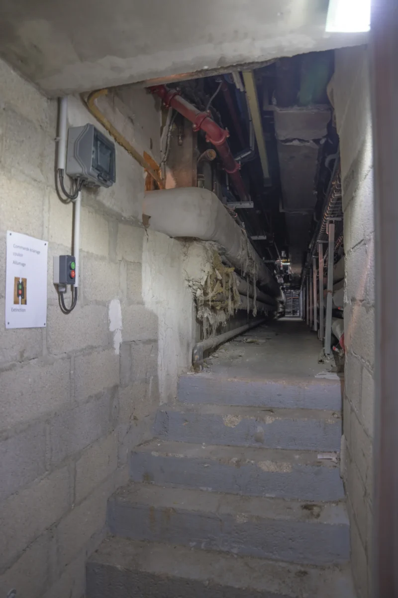 Escalier étroit dans un sous-sol technique avec câbles, tuyaux et équipements électriques apparents, ambiance industrielle