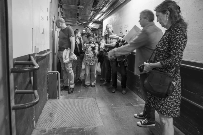Groupe de personnes debout dans un espace étroit, probablement un navire ou un lieu historique, observant ou discutant.