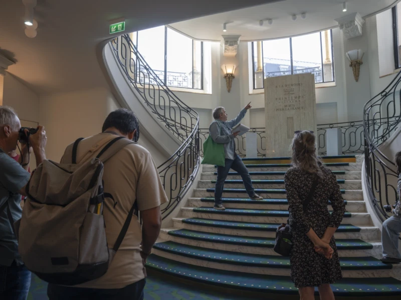 Alice · 20/09/2026 · 3840 x 2560 Cette photographie montre un groupe de personnes dans un escalier intérieur. Le sujet principal est une femme en vert, gesticulant comme pour expliquer ou guider le groupe. La composition met en valeur les escaliers en colimaçon et les murs clairs. Le cadrage est serré, centré sur les personnes et l'escalier.La lumière est naturelle et douce, éclairant uniformément la scène. Les couleurs dominantes sont les tons neutres (beige, gris) avec des touches de vert et de noir. Le style est documentaire, capturant un moment réel. L'ambiance est informative et conviviale, évoquant une visite ou une présentation guidée.