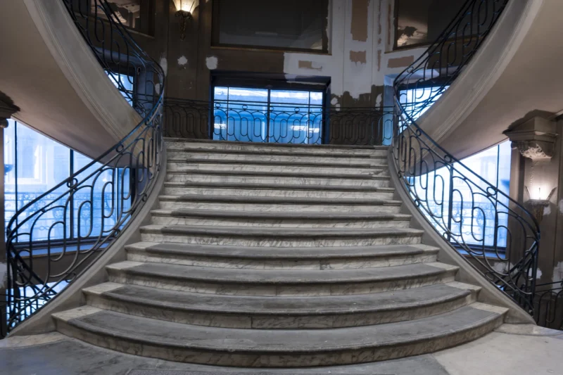 Escalier monumental en marbre avec balustrades en fer forgé bleuâtre, vue symétrique depuis le bas vers un palier supérieur