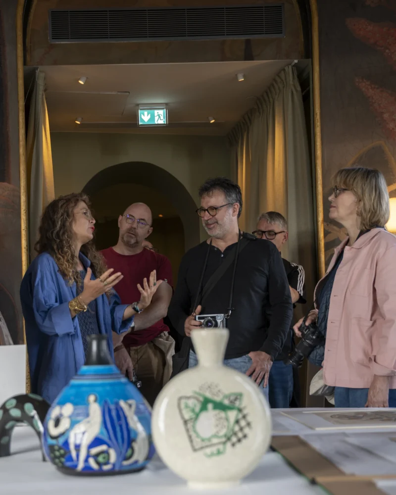 Groupe de visiteurs écoutant une guide dans une exposition d'art avec des vases anciens en vitrine sous une lumière tamisée