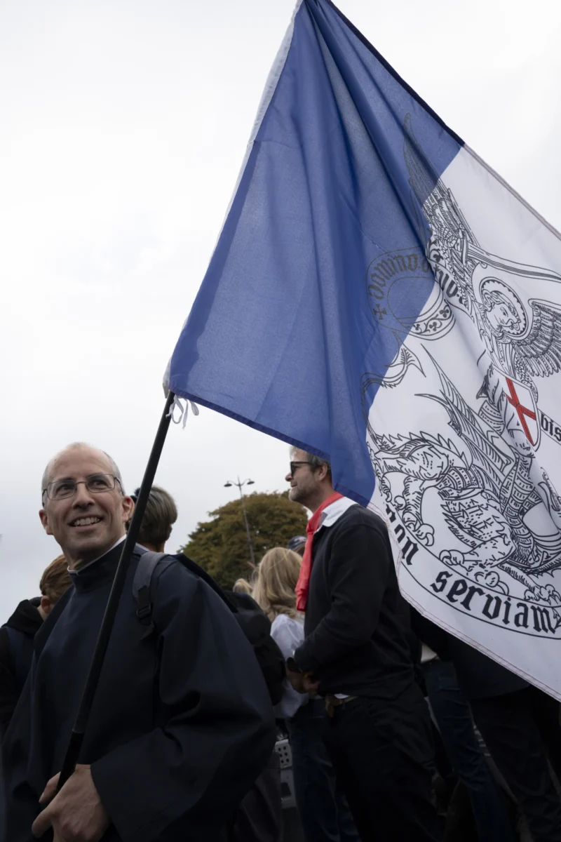 Alice · 26/09/2026 · 2560 x 3840 Manifestation avec une bannière aux emblèmes religieux et texte latin, plusieurs personnes marchant en extérieur sous