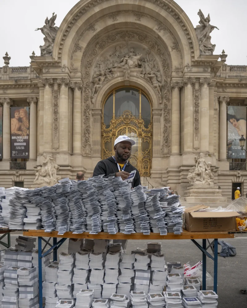 Alice · 26/09/2026 · 3072 x 3840 Un vendeur de livres devant un bâtiment orné de sculptures et de dorures, avec des piles de livres exposées sur une table.