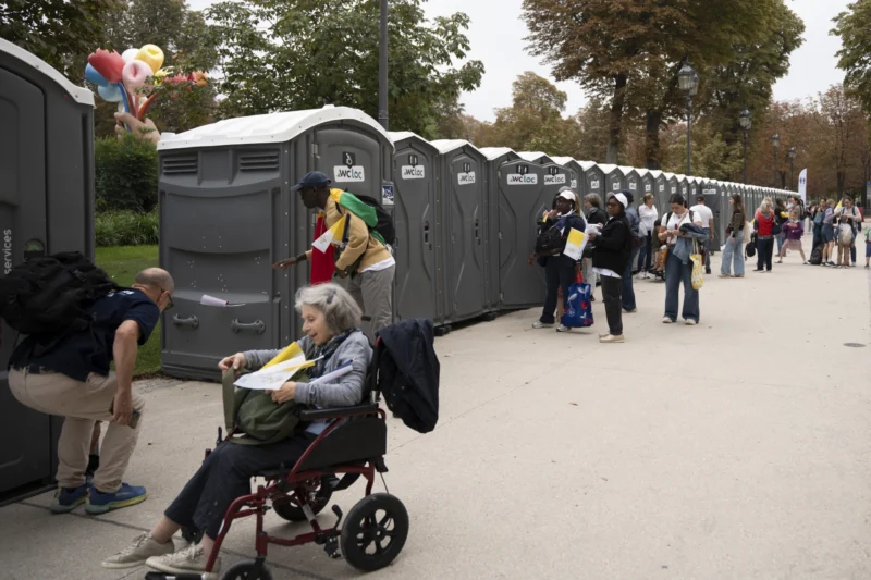 Alice · 26/09/2026 · 3840 x 2560 Une personne en fauteuil roulant attend devant une file de toilettes portables dans un parc, avec d'autres personnes
