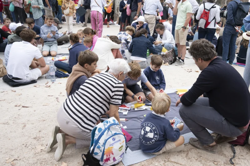 Alice · 26/09/2026 · 3840 x 2560 Groupe d'enfants et d'adultes assis sur une surface plane en extérieur, occupés à dessiner ou jouer avec des feuilles et