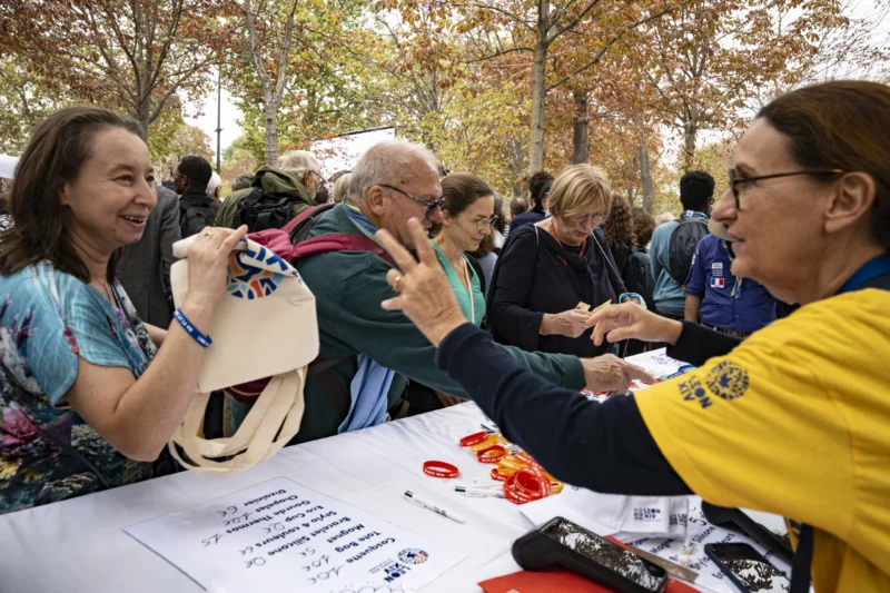 Alice · 26/09/2026 · 3840 x 2560 Un groupe de personnes participant à un événement en extérieur, échangeant des objets et des sourires autour d'une table