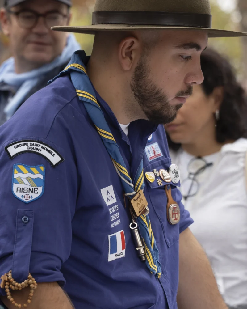 Alice · 26/09/2026 · 3072 x 3840 Un individu en tenue de scout avec insignes et écharpe, participant à une cérémonie ou rassemblement en extérieur.