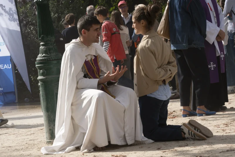 Alice · 26/09/2026 · 3840 x 2560 Un individu vêtu de blanc avec une cape et une robe colorée discutant avec une personne en tenue décontractée près d'un