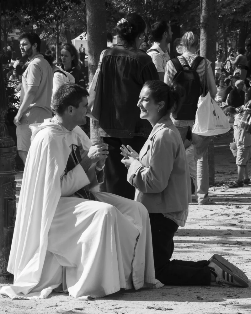 Alice · 26/09/2026 · 3072 x 3840 Deux personnes assises en conversation dans un cadre urbain entouré d'une foule, l'une vêtue de blanc, l'autre en tenue