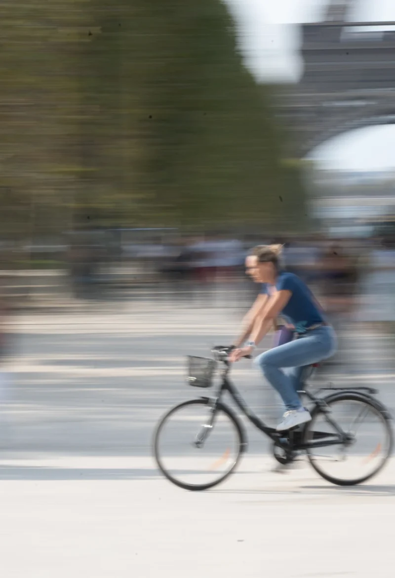 Jean-Marc L. · 26/09/2026 · 1559 x 2284 Cycliste en mouvement sur une piste, vêtue d'un t-shirt bleu et de jeans, avec un flou artistique suggérant la vitesse.