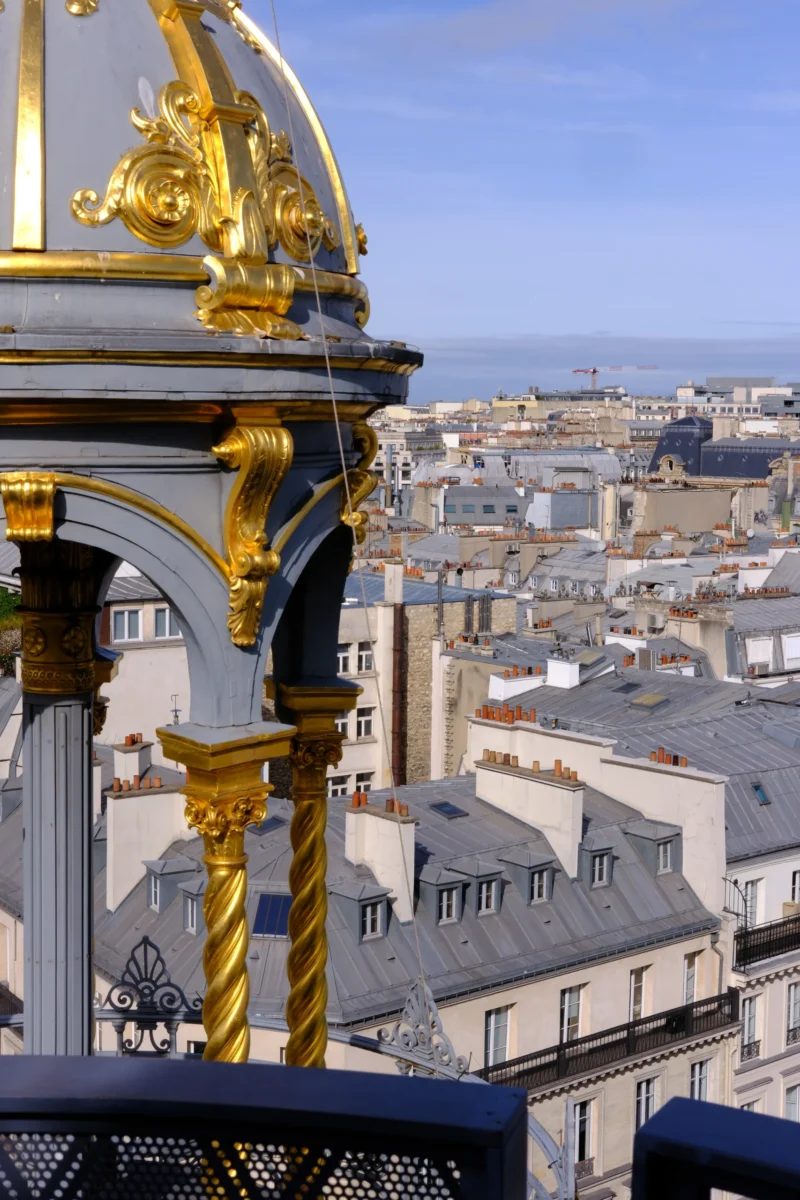 Vue depuis une balustrade dorée sur des toits parisiens aux tuiles grises et bâtiments classiques sous un ciel dégagé