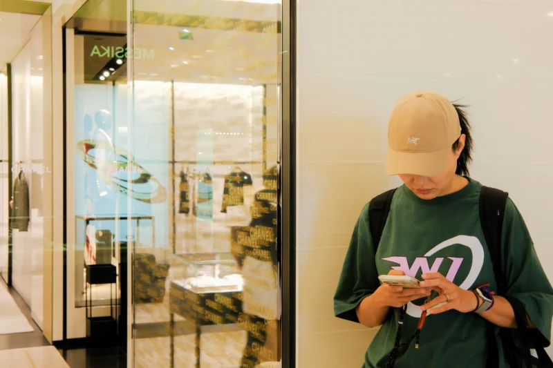 Une personne en capuche verte et casquette beige utilise son téléphone portable près d’une vitrine de magasin reflétant