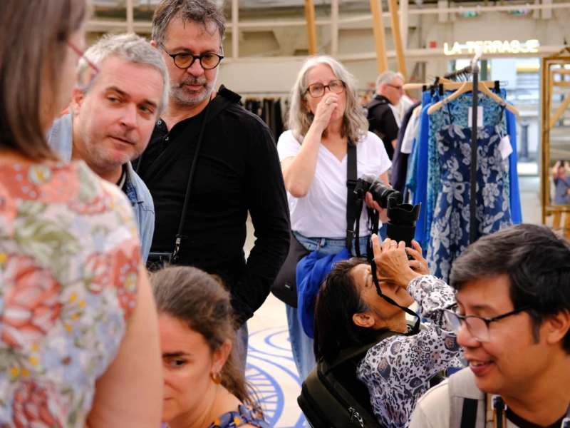 Nathalie F. · 20/09/2026 · 7728 x 5152 Cette photographie montre un groupe de personnes dans un magasin de vêtements. Le sujet principal semble être une interaction entre les individus, notamment une personne prenant une photo avec un smartphone.La composition est serrée, centrée sur les visages et les interactions. Le cadrage est en plan moyen, mettant en valeur les expressions et les détails vestimentaires. La lumière est naturelle et douce, éclairant uniformément la scène. Les couleurs dominantes sont neutres et pastel, avec des touches de couleurs vives sur les vêtements.L’ambiance est conviviale et informelle, évoquant une sortie entre amis ou une activité sociale.