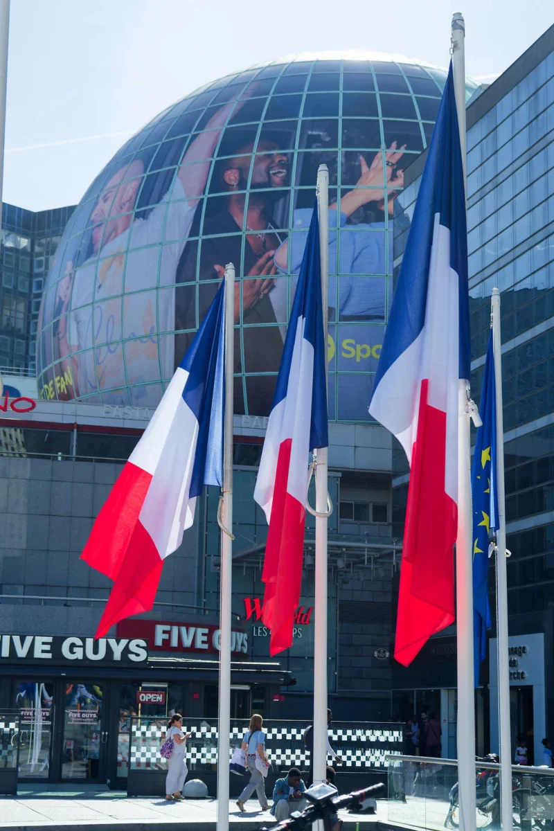 Monique · 05/09/2026 · 1280 x 1920 Drapeaux français flottant devant un bâtiment moderne avec une façade reflétant des visages et des publicités, devant