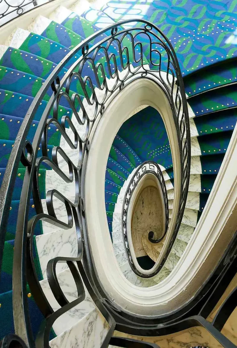 Escalier en colimaçon avec balustrade en fer forgé et revêtement mural coloré aux tons bleus et verts, vue depuis le bas