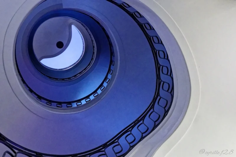 Vue plongeante d'un escalier en colimaçon avec des marches bleutées et une rampe en métal, évoquant un espace architectural