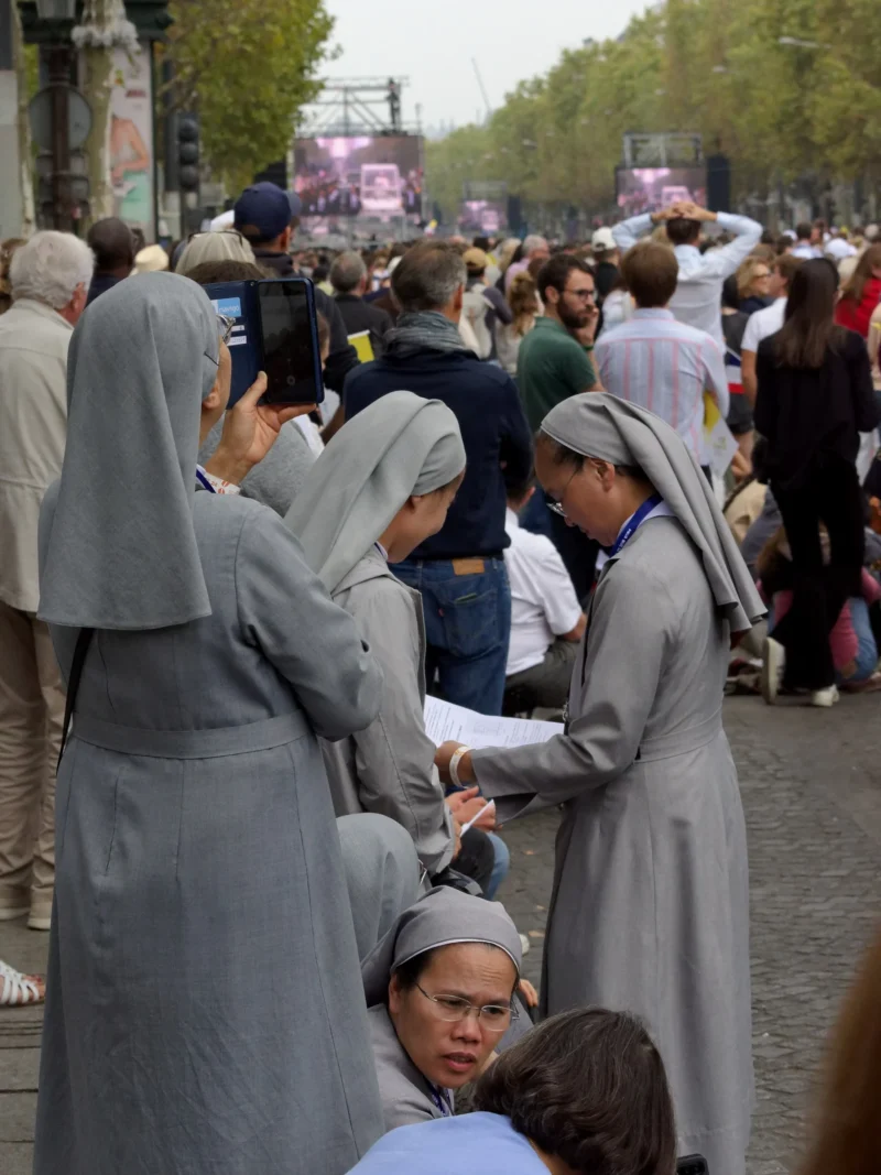 Cyrille · 26/09/2026 · 1519 x 2026 Quatre religieuses en habits gris distribuant des documents ou objets à des personnes assises ou debout lors d'une