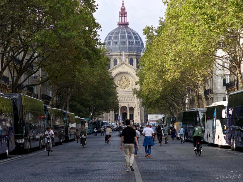 Cyrille · 26/09/2026 · 2026 x 1520 Rue animée bordée d'arbres menant à un imposant bâtiment avec une horloge et une coupole, sous un ciel clair, avec des bus
