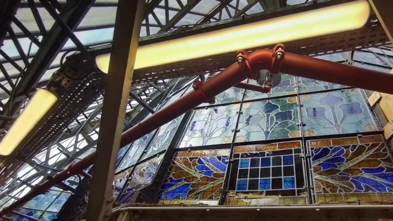 Vitrail coloré représentant des motifs floraux et géométriques dans une structure métallique éclairée par des néons jaunes.