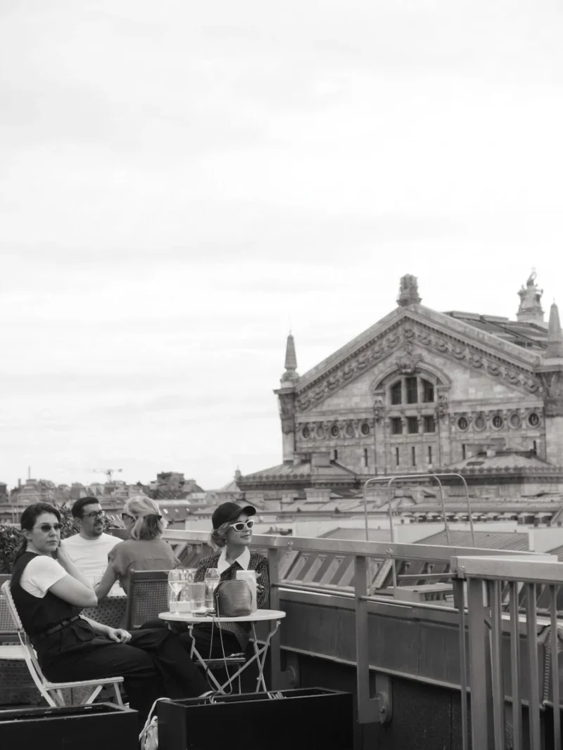 Quatre personnes assises sur une terrasse en ville avec une vue sur un bâtiment historique en arrière-plan, en noir et blanc