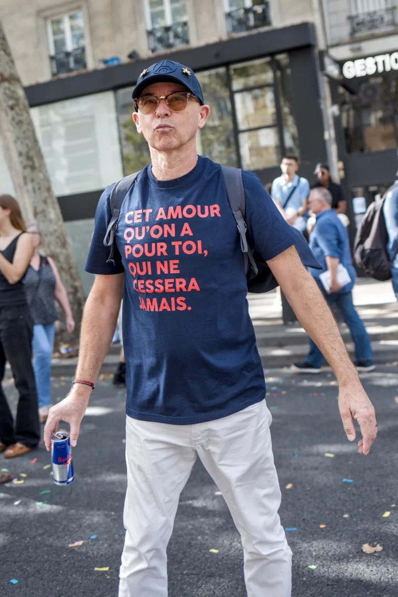 Frédéric · 19/09/2026 · 1120 x 1680 Un homme en t-shirt à message en français, casquette et pantalon blanc, tenant une canette, dans une rue animée avec