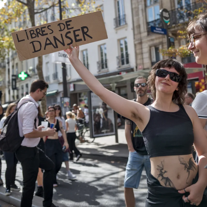 Frédéric · 19/09/2026 · 1680 x 1680 Manifestante levant une pancarte en carton disant 'Libres de danser' dans une rue animée, entourée de passants.