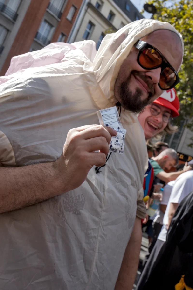 Frédéric · 19/09/2026 · 1120 x 1680 Un homme en combinaison blanche tenant une carte ou un badge devant un groupe de personnes en extérieur, sourire visible.