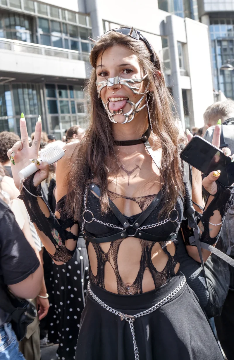 Frédéric · 19/09/2026 · 1094 x 1680 Personne en costume gothique et accessoires métalliques posant en public avec un geste de la main, entourée d'autres