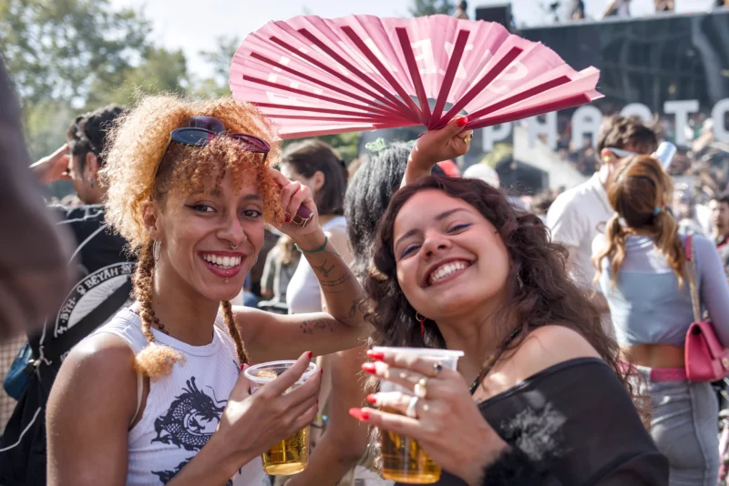Frédéric · 19/09/2026 · 2517 x 1680 Deux femmes souriantes en fête tenant des verres et un éventail, entourées d’une foule animée en extérieur sous un ciel clair