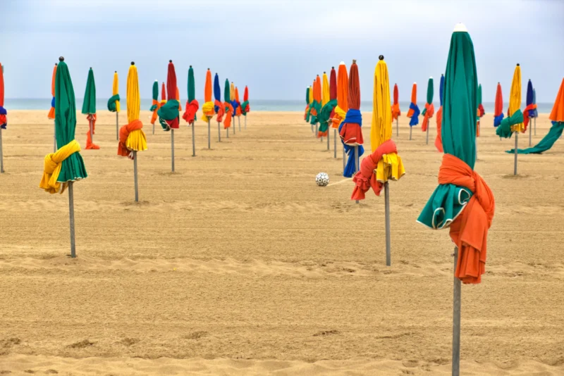 Fabienne · 17/07/2026 · 5496 x 3670 Plage déserte avec rangées dombrelles colorées alignées sur le sable, sous un ciel couvert.