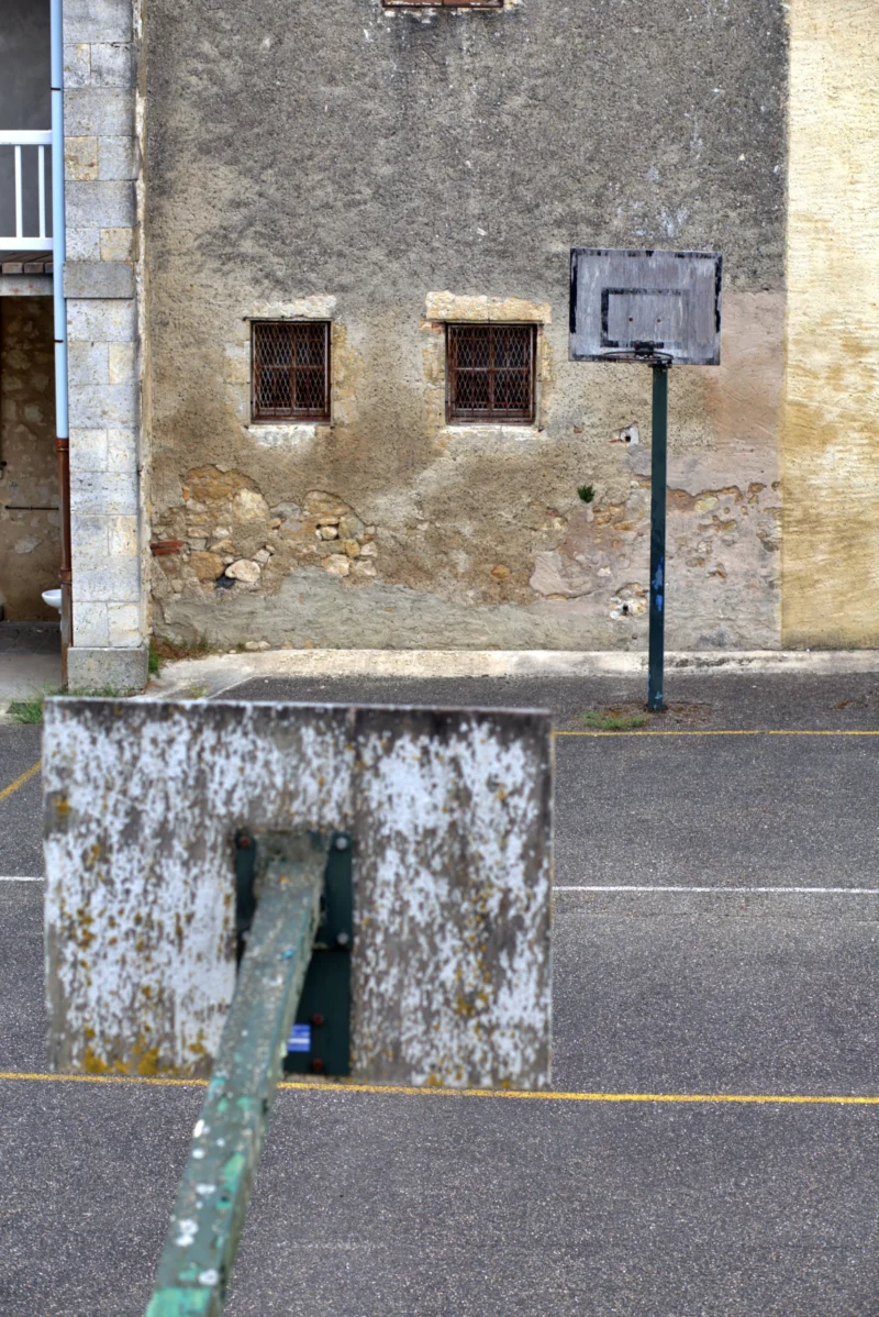 Fabienne · 31/07/2026 · 3670 x 5496 Ancien bâtiment en pierre avec une façade abîmée, deux fenêtres grillagées et un panneau de basket fixé sur un poteau vert