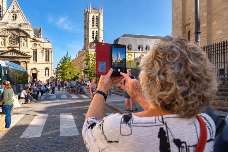 Bao · 12/09/2026 · 1536 x 1024 Une personne prend une photo d’une cathédrale gothique avec un smartphone en plein jour, entourée de passants dans une place