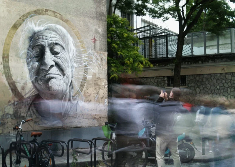 Bao · 5386 x 3816 Murale d'un visage âgé stylisé sur un mur de ville avec des passants floutés en mouvement devant des bicyclettes et