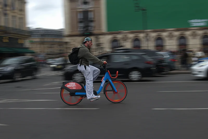 Bao · 04/06/2026 · 6024 x 4016 Un cycliste en mouvement sur un vélo électrique rouge et bleu dans une rue animée avec des voitures floues en arrière-plan