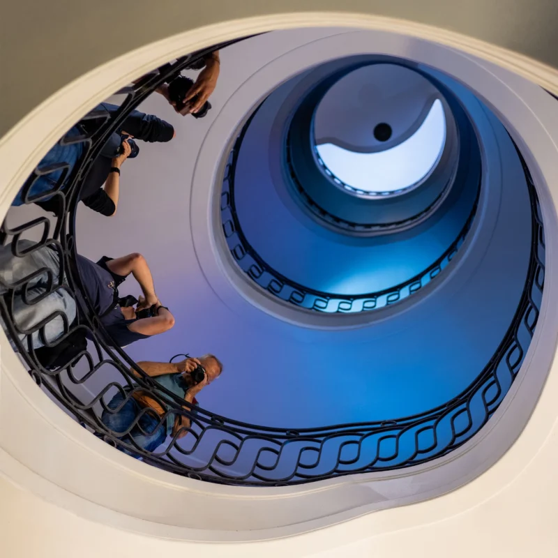 Vue plongeante d'un escalier en colimaçon bleu éclairé, avec des personnes descendant et tenant des sacs, ambiance