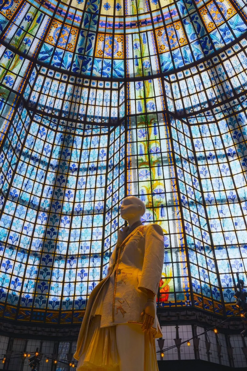 Statue dorée sous une verrière aux vitraux colorés représentant des motifs floraux et géométriques, vue de dessous vers