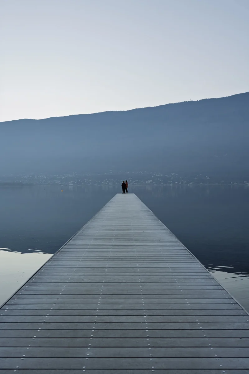 Cécile · 29/12/2025 · 1280 x 1920 Personne debout au bout d'une longue jetée en bois sur un lac calme, entourée de montagnes à l'horizon.