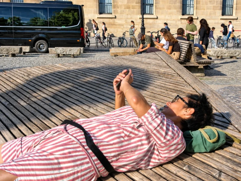 Bao · 12/09/2026 · 1432 x 1098 Cette photographie montre une personne allongée sur un banc en bois, sujet principal, semblant utiliser son téléphone. La composition est simple et naturelle, avec un cadrage serré sur le sujet, laissant en arrière-plan des passants et des vélos.La lumière est vive et ensoleillée, créant des ombres marquées. Les couleurs dominantes sont les tons chauds : rouge et blanc des rayures du vêtement, vert de la veste, et les teintes neutres du bois et des pierres. Le style est documentaire et spontané, évoquant une ambiance détendue et urbaine, typique d’une journée ensoleillée en ville.
