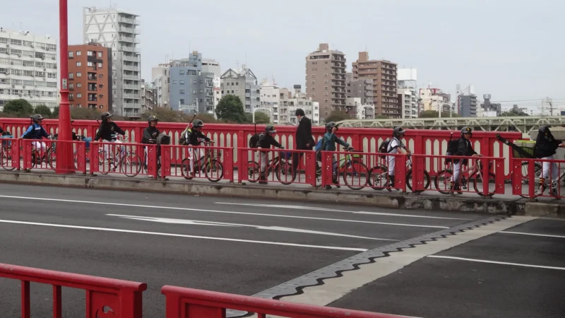 Bao · 30/10/2016 · 1920 x 1080 Un groupe de cyclistes roulant sur un pont rouge en ville, avec des immeubles en arrière-plan.