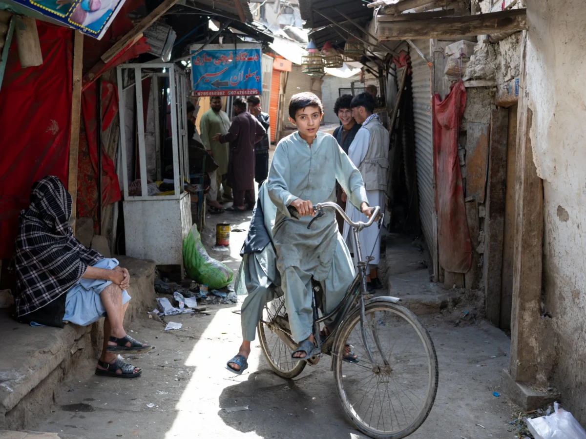 Daniel · 27/05/2026 · 2400 x 1800 Un jeune homme sur un vélo dans une ruelle animée entourée de petits commerces et de personnes vaquant à leurs occupations.