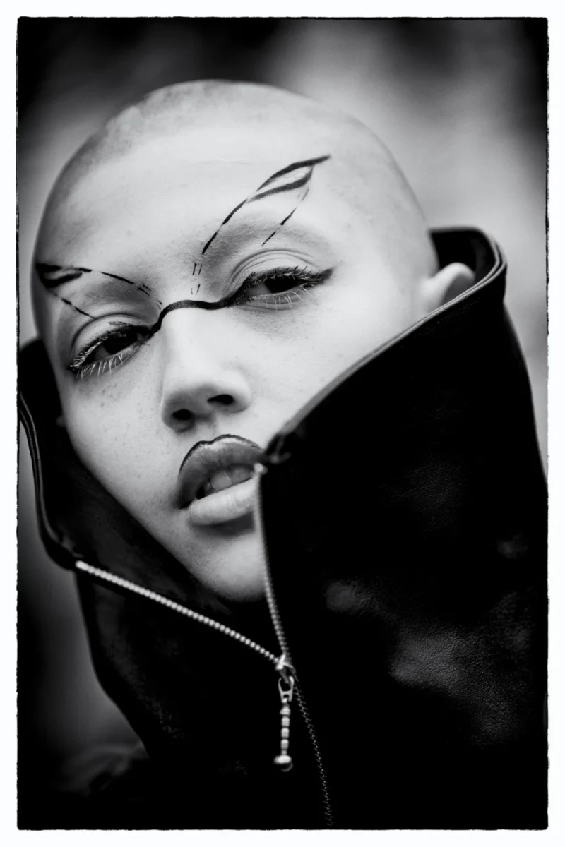 Portrait stylisé d’un visage aux traits marqués et maquillage futuriste, coiffé d’un bonnet, tenant un accessoire devant