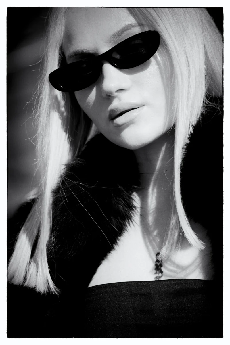 Portrait en noir et blanc d’une personne aux cheveux longs portant des lunettes et une veste à fourrure, regard légèrement
