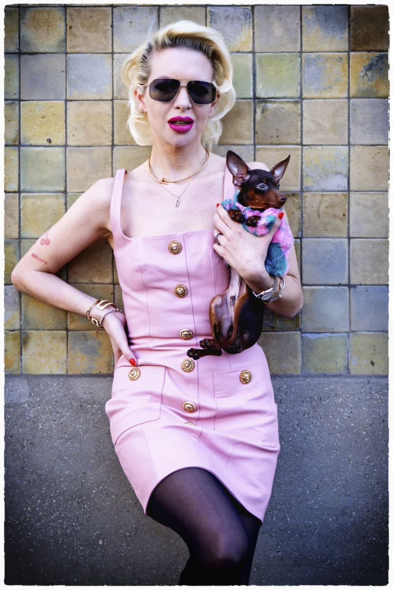 Femme stylisée en tenue élégante rose avec boutons dorés, posant avec un petit chien habillé contre un mur en carreaux
